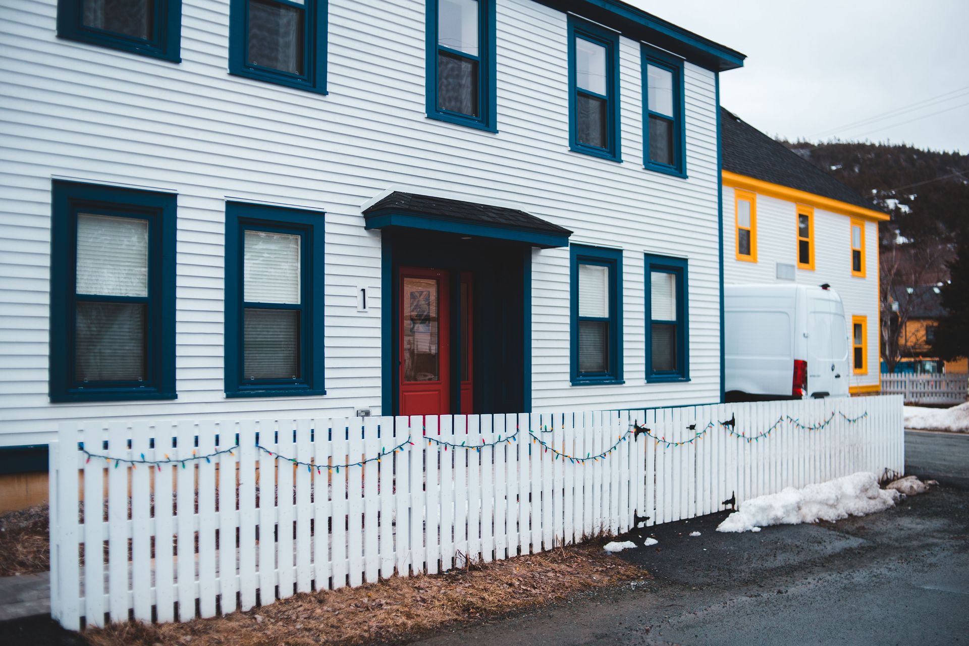 A charming residential house with a white fence, adorned with string lights and snowy surroundings.